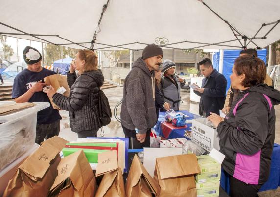 People talking to each other under a canopy. There are paper bags and other supplies on the tables.