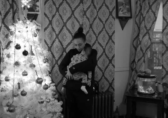 An individual is carrying a baby standing next to a Christmas tree.