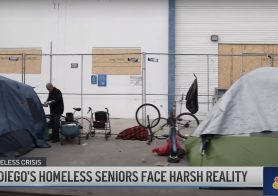 Individuals experiencing homelessness are near tents next to a white building.