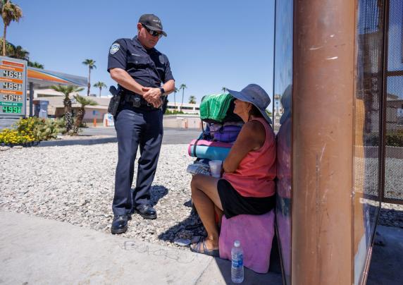  Photo of police officer speaking with homeless woman sitting in the shade of a bus stop