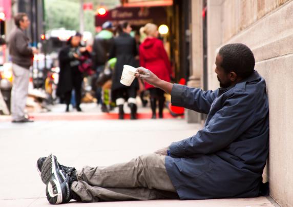 Photo of a man sitting on the sidewalk holding up a cup